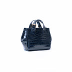 A small structured handbag with a glossy crocodile-embossed texture in black, featuring dual top handles and a gold-tone logo plate on the front, set against a plain white background.