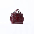 A small structured handbag with a glossy crocodile-embossed texture in deep burgundy, featuring dual top handles and a top zipper closure, set against a plain white background.