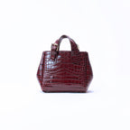 A small structured handbag with a glossy crocodile-embossed texture in deep burgundy, featuring dual top handles and a gold-tone buckle detail, set against a plain white background.