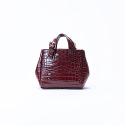 A small structured handbag with a glossy crocodile-embossed texture in deep burgundy, featuring dual top handles and a gold-tone buckle detail, set against a plain white background.