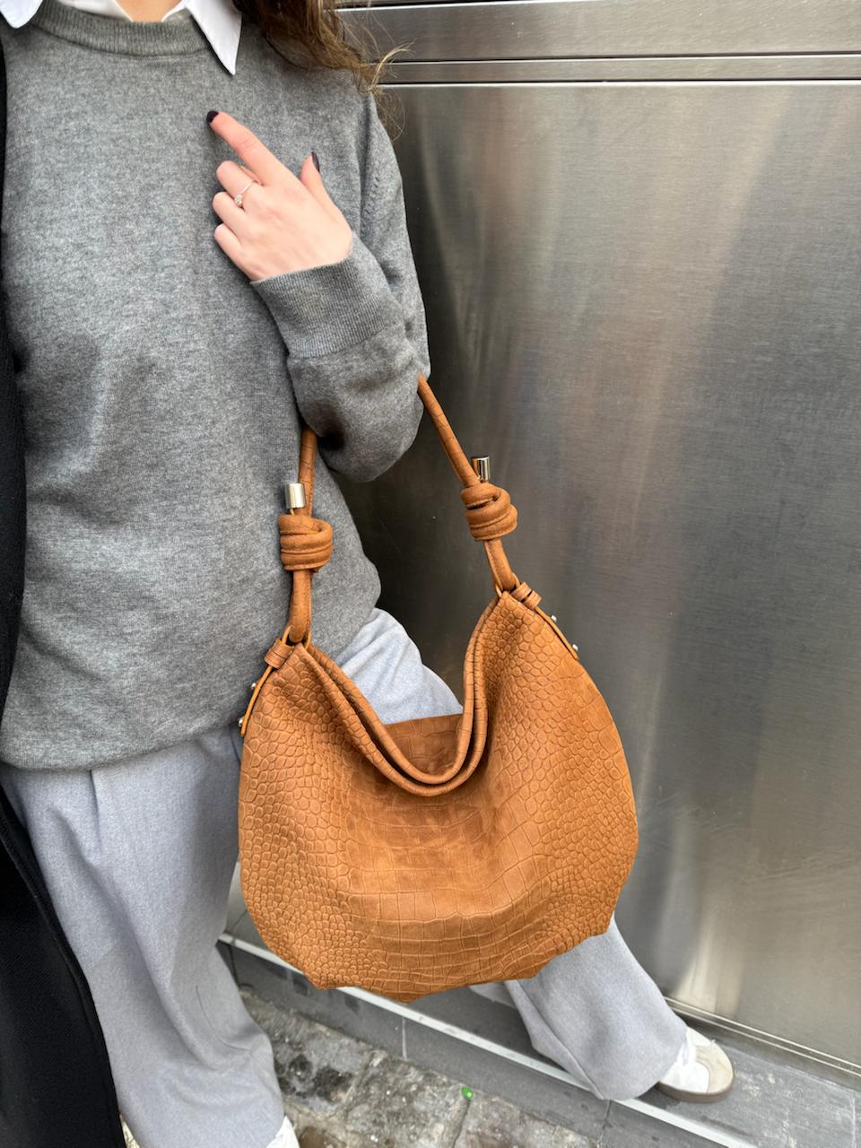 A person wearing a gray sweater and light gray sweatpants carries a tan pebbled leather hobo shoulder bag with knotted strap details against a metallic gray wall.