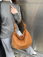 A person wearing a gray sweater and light gray sweatpants carries a tan pebbled leather hobo shoulder bag with knotted strap details against a metallic gray wall.